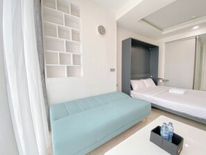 A modern hotel room with a light blue sofa, a bed with white linens, a small table with water bottles, and a wall-mounted TV. - Luxurious Studio At Art Deco Apartment (Bandung)