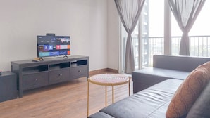 A modern living room with a flat-screen TV on a dark wooden TV stand, a round wooden side table, and a grey sofa with a decorative pillow. - Cozy Living 2Br Apartment At Casa De Parco (Pagedangan)