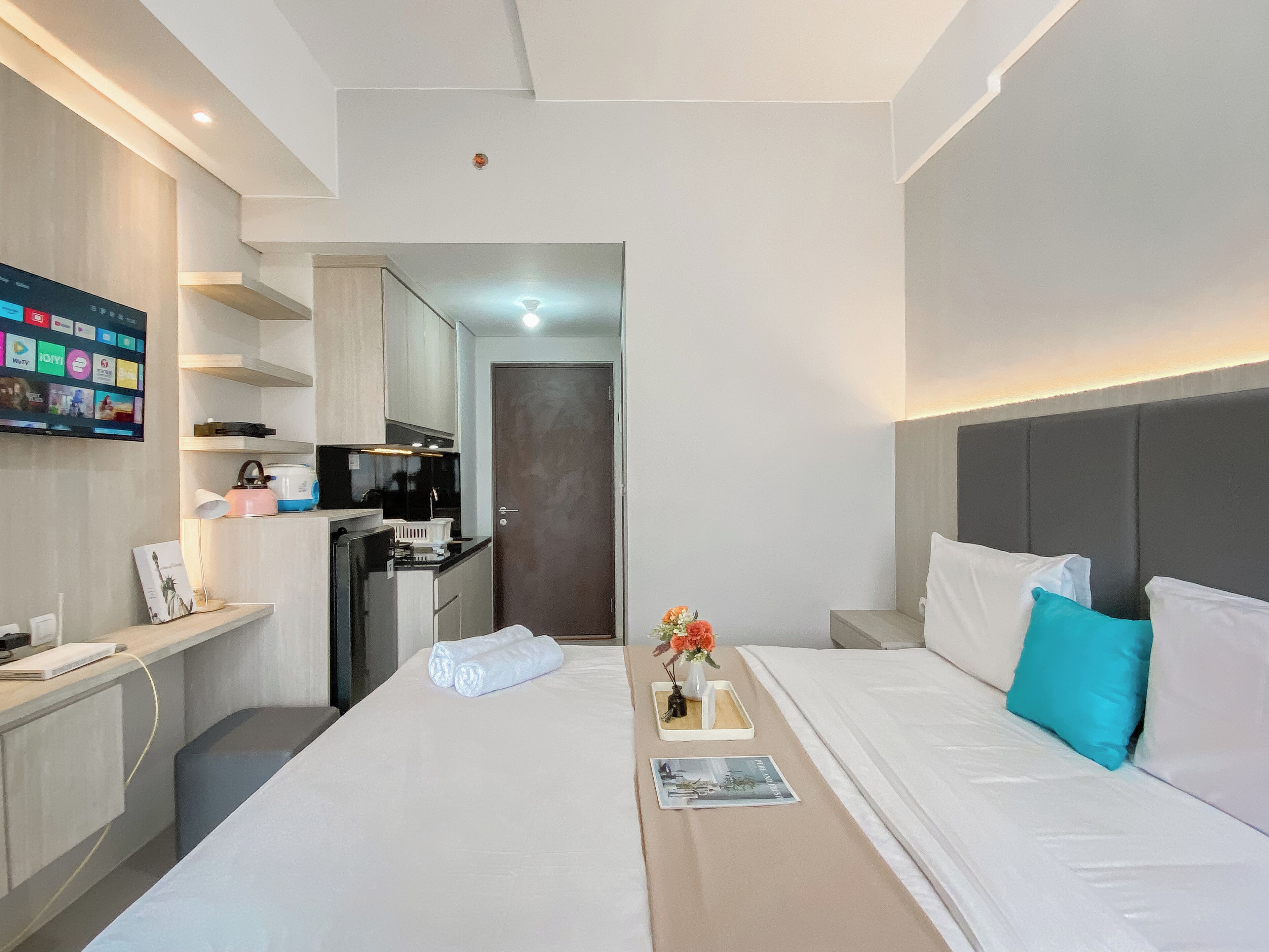 A modern hotel room with a bed, bedside tables, a small desk, a flat-screen TV, a kitchenette, and a bathroom.