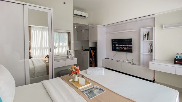 A hotel room with a bed, a bedside table with a vase of flowers, a flat-screen TV mounted on the wall, and a small kitchenette. - Good Place And Comfortable Studio Apartment Springlake Summarecon Bekasi (Bekasi)