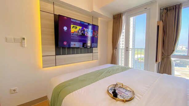 Een hotelkamer met een groot bed, een wandtelevisie waarop verschillende zenders worden getoond, een raam met gordijnen en uitzicht op de stad.
