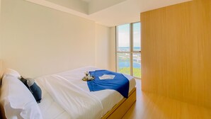 A modern hotel room with a large bed, a wooden wardrobe, and a view of a cityscape through a large window. - Good Place And Modern 1Br Gold Coast Apartment (Jakarta)
