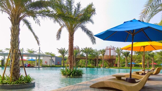 A poolside area with lounge chairs and umbrellas, surrounded by palm trees and a water slide structure. - Good Place And Modern 1Br Gold Coast Apartment (Jakarta)