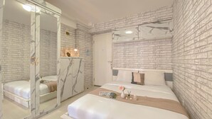 A bedroom with a marble-framed mirror, a brick-patterned wall, a bed with white bedding, and a bedside table with a vase and candles. - Homey And Well Furnished Studio At Bassura City Apartment (Jakarta)