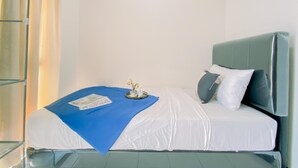 Een netjes opgemaakt bed met een blauwe deken, witte lakens en een kussen. Er staat een klein tafeltje met een vaas en een boek naast het bed.