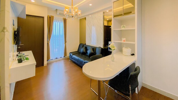 A modern living room with a sofa, a dining table with chairs, a chandelier, and a large window with curtains. - Good And Warm 2Br At Transpark Cibubur Apartment (Depok)