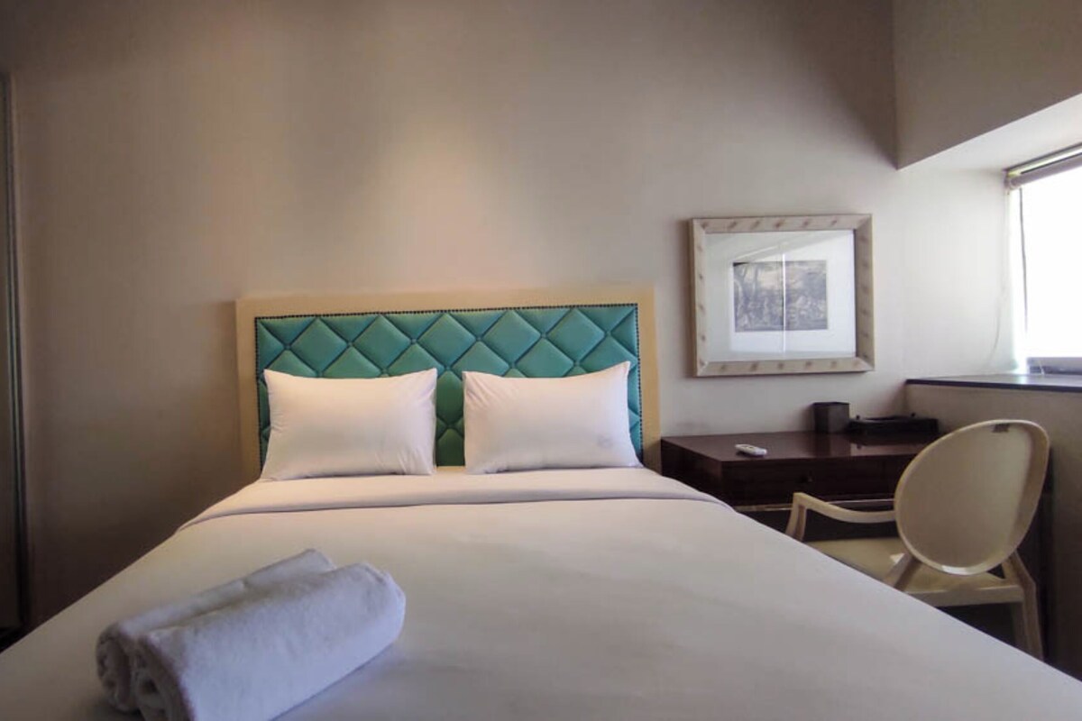 A hotel room with a large bed, a headboard with a diamond pattern, two white pillows, a neatly folded white towel, a desk with a chair, and a framed picture on the wall.