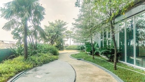 A curved pathway lined with palm trees and other greenery, leading to a modern building with large windows. - Spacious And Homey 2Br Apartment With Private Lift At The Kencana Residence (Jakarta)