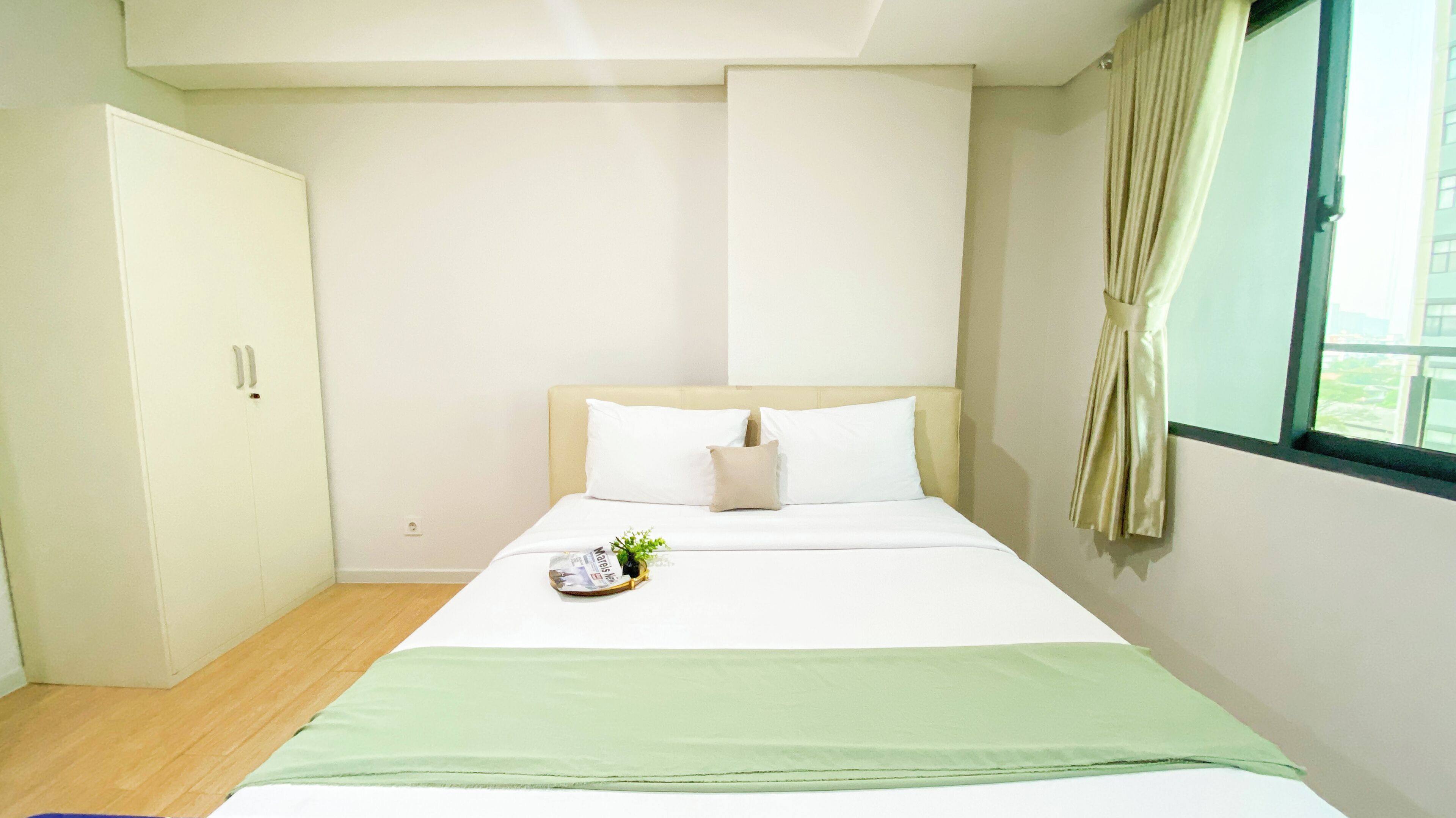 A hotel room with a bed, a green throw blanket, a small plant, and a window with curtains.