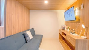 A room with a blue sofa, wooden panel walls, a TV mounted on the wall, and a wooden cabinet with various items. - Studio At Transpark Juanda Bekasi Timur Apartment (Bekasi)