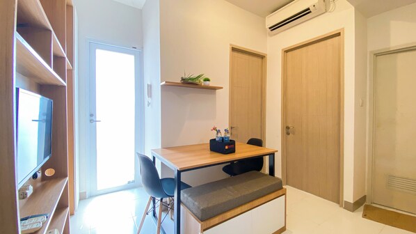 Een kleine, goed verlichte kamer met een houten bureau, een bankje, een stoel en een plank met planten. Er is een deur met een raam en een airconditioner aan de muur.