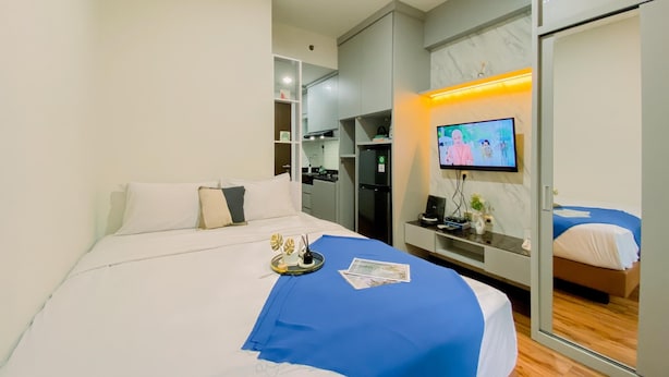 A modern hotel room with a bed, a desk, a television, and a kitchen area. - Cozy Studio Apartment At Transpark Cibubur (Depok)