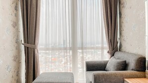 Ein modernes Wohnzimmer mit einem grauen Sofa, dazu passendem Hocker und einem Holztisch. Das Zimmer zeichnet sich durch durchscheinende Vorhänge und blumenmusterierte Tapeten aus.