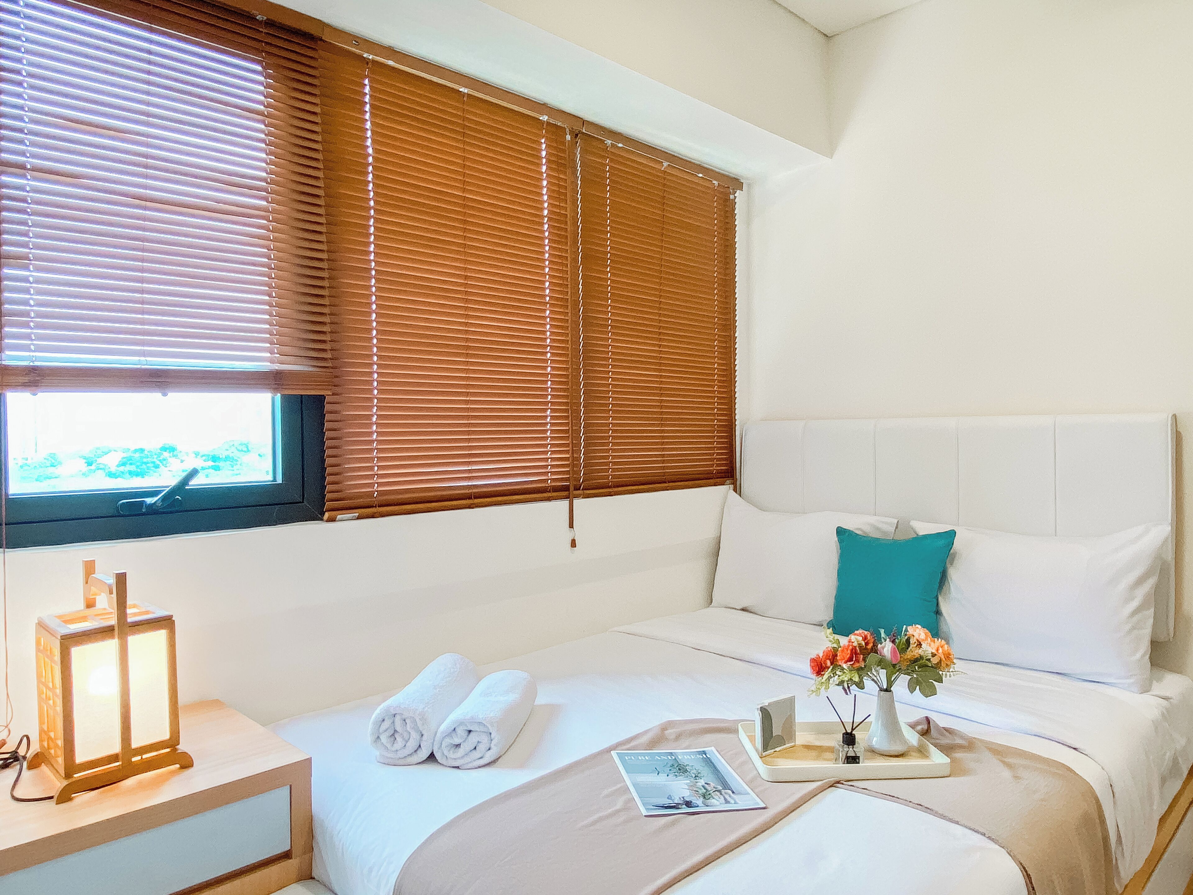 A hotel room with a bed, pillows, a nightstand, a lamp, and a window with blinds.