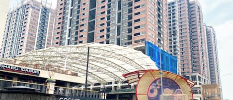 Una calle urbana moderna con edificios altos, un área de estar al aire libre cubierta y diversos establecimientos comerciales, incluyendo una cafetería.