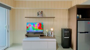 A living room with a flat-screen TV on a white cabinet, a refrigerator, and a wooden shelf above the TV.