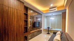 A modern living room with a wooden cabinet, a flat-screen TV mounted on a marble wall, a sofa with cushions, and a small table with a bottle. - Well Furnished 1Br At Anderson Supermall Mansion Apartment (Surabaya)