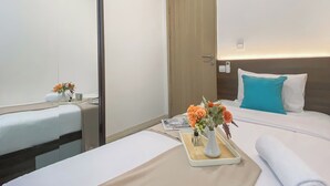 A hotel room with a bed, a bedside table with a vase of flowers, a mirror, a wooden door, and a wall-mounted air conditioning unit. - Homey And Modern Look 3Br At Meikarta With Balcon Apartment (Cikarang)