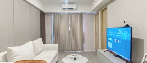 A modern hotel room with a white sofa, a small round table, a TV on a stand, and a curtain in the background.