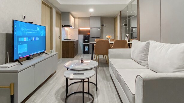 A modern living room with a flat-screen TV, a white sofa, a coffee table, and a dining area with chairs and a table. - Homey And Modern Look 3Br At Meikarta With Balcon Apartment (Cikarang)