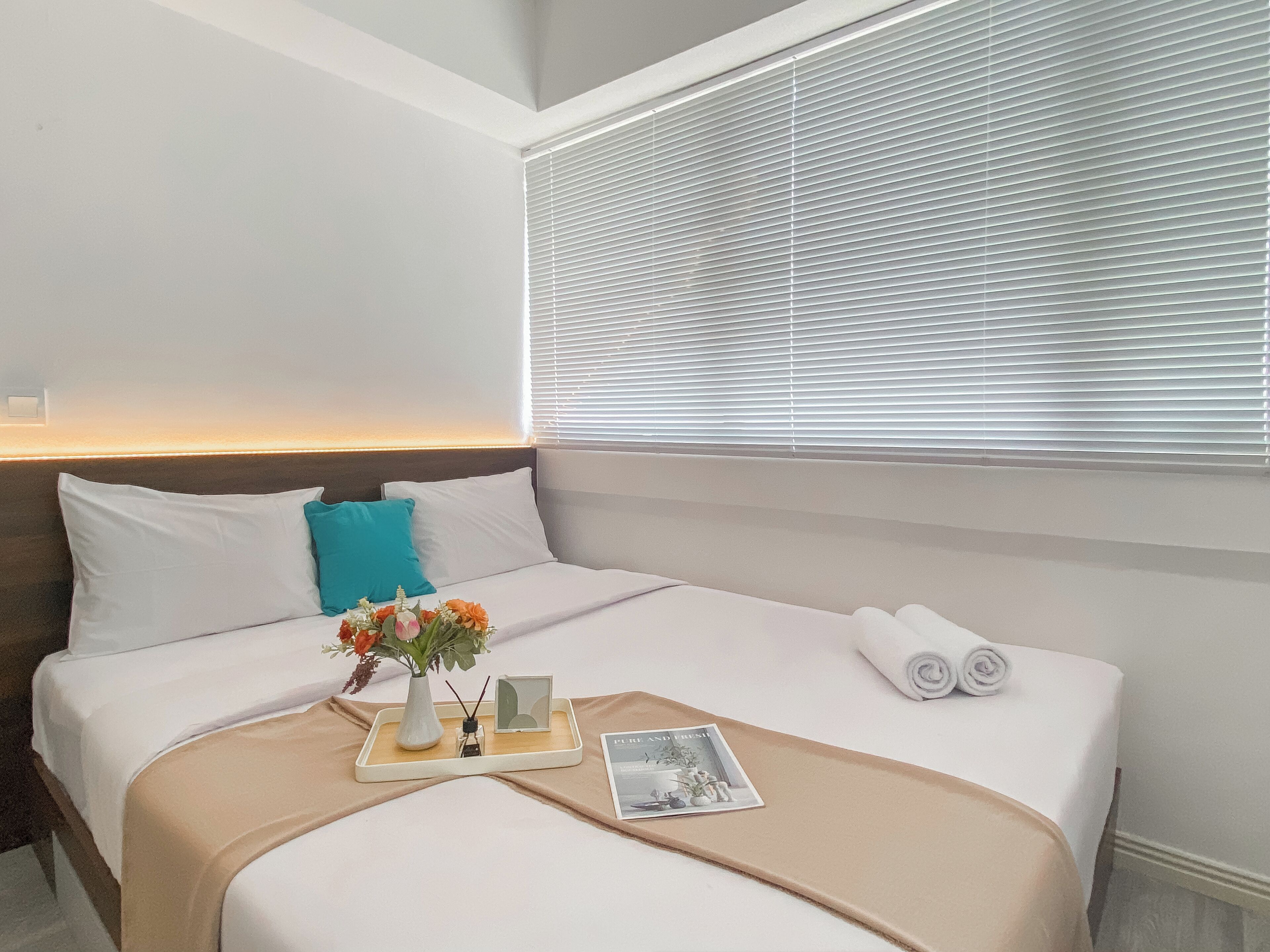 A neatly made bed with white and teal pillows, a vase with flowers, a tray with a book and a picture, and a window with blinds.