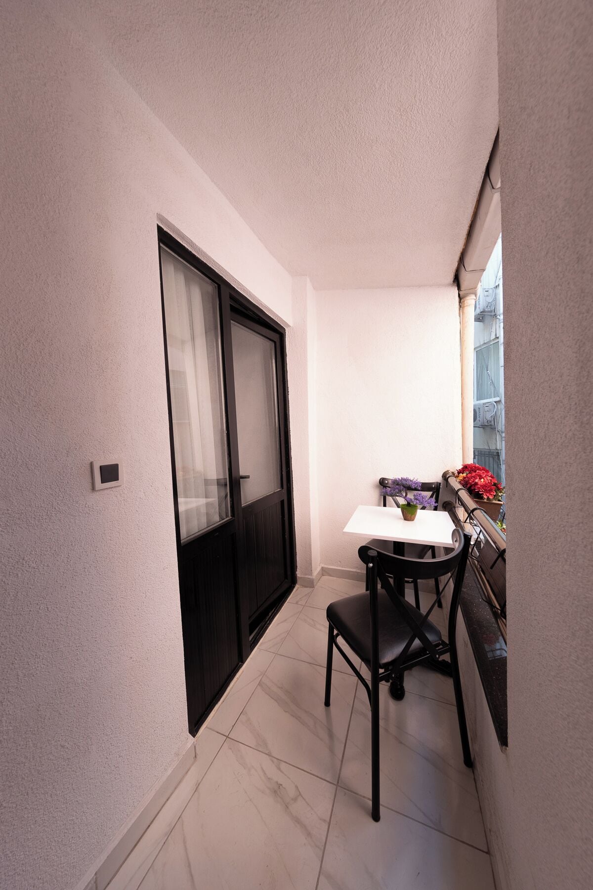 A small balcony with a white table and two chairs, a sliding glass door, and potted plants.