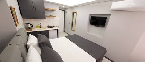 A compact room with a bed, a kitchenette, a TV, and an air conditioning unit.