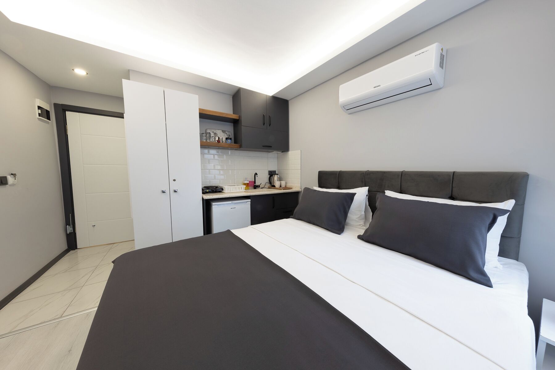 A modern bedroom with a bed, bedside tables, a built-in wardrobe, a kitchen area with a sink and countertop, and an air conditioning unit.