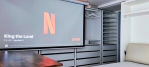 Quarto moderno com uma grande TV de tela plana exibindo 'King the Land' na Netflix, um guarda-roupa com espaço para pendurar roupas e uma cama com cabeceira bege.