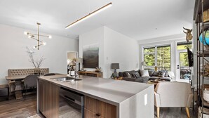 A modern kitchen with a central island, a dining area with a sofa, and a living room with a large window. - Beautiful & Modern Condo, Ski-In Ski-Out (Sun Peaks)