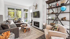 A modern living room with a sectional sofa, a coffee table, and a fireplace. There is a bookshelf with decorative items and a mounted TV. - Beautiful & Modern Condo, Ski-In Ski-Out (Sun Peaks)
