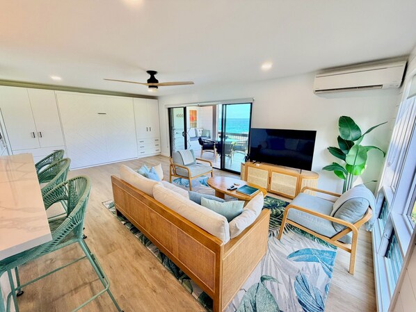 A modern living room with a sofa, chairs, a coffee table, and a TV. There is a large window with a view of the ocean.