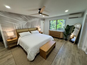 A modern bedroom with a large bed, wooden nightstands, a bench, a dresser, a ceiling fan, and a window with a view of greenery.
