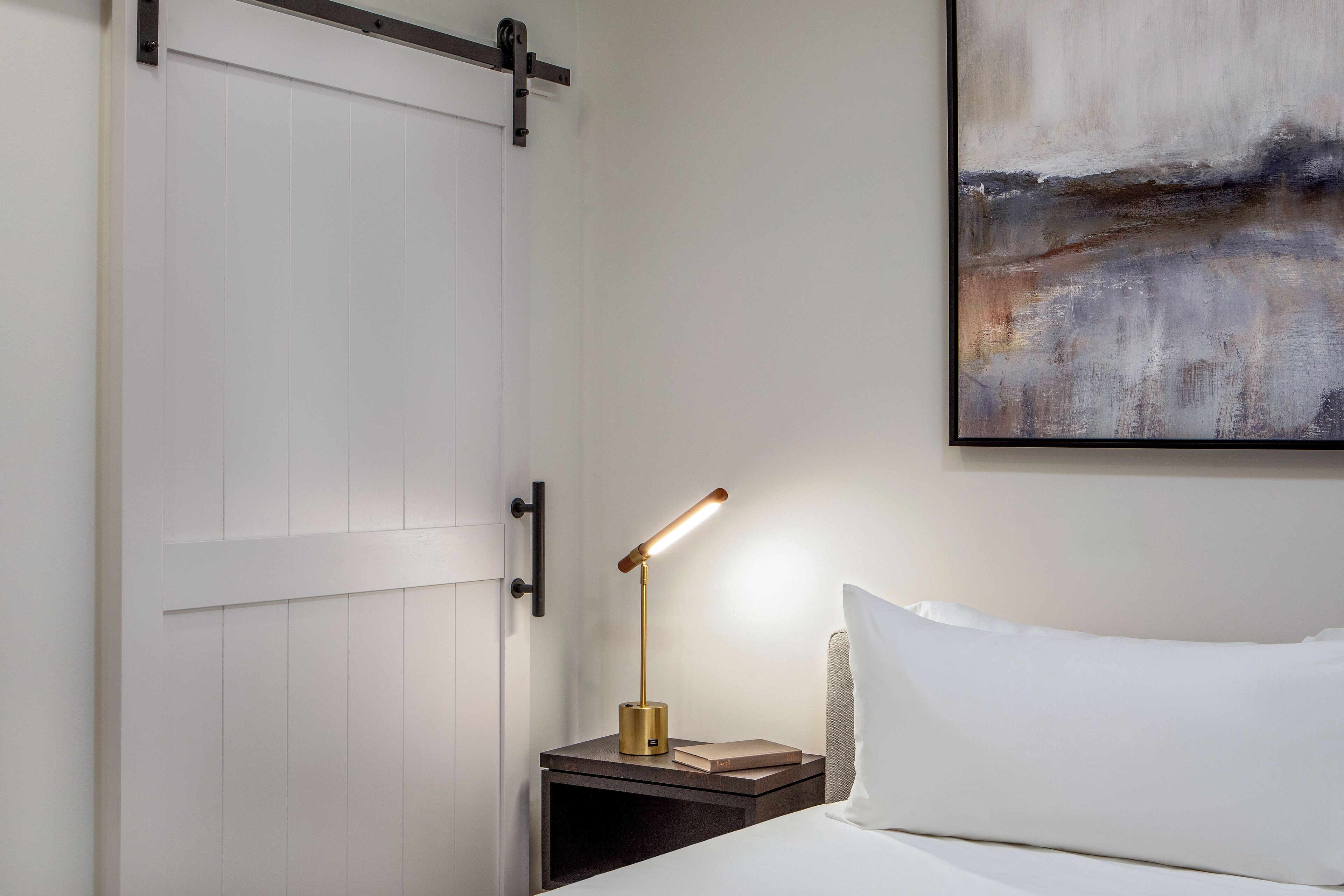 A neatly made bed with white bedding, a bedside table with a lamp, and a painting on the wall.