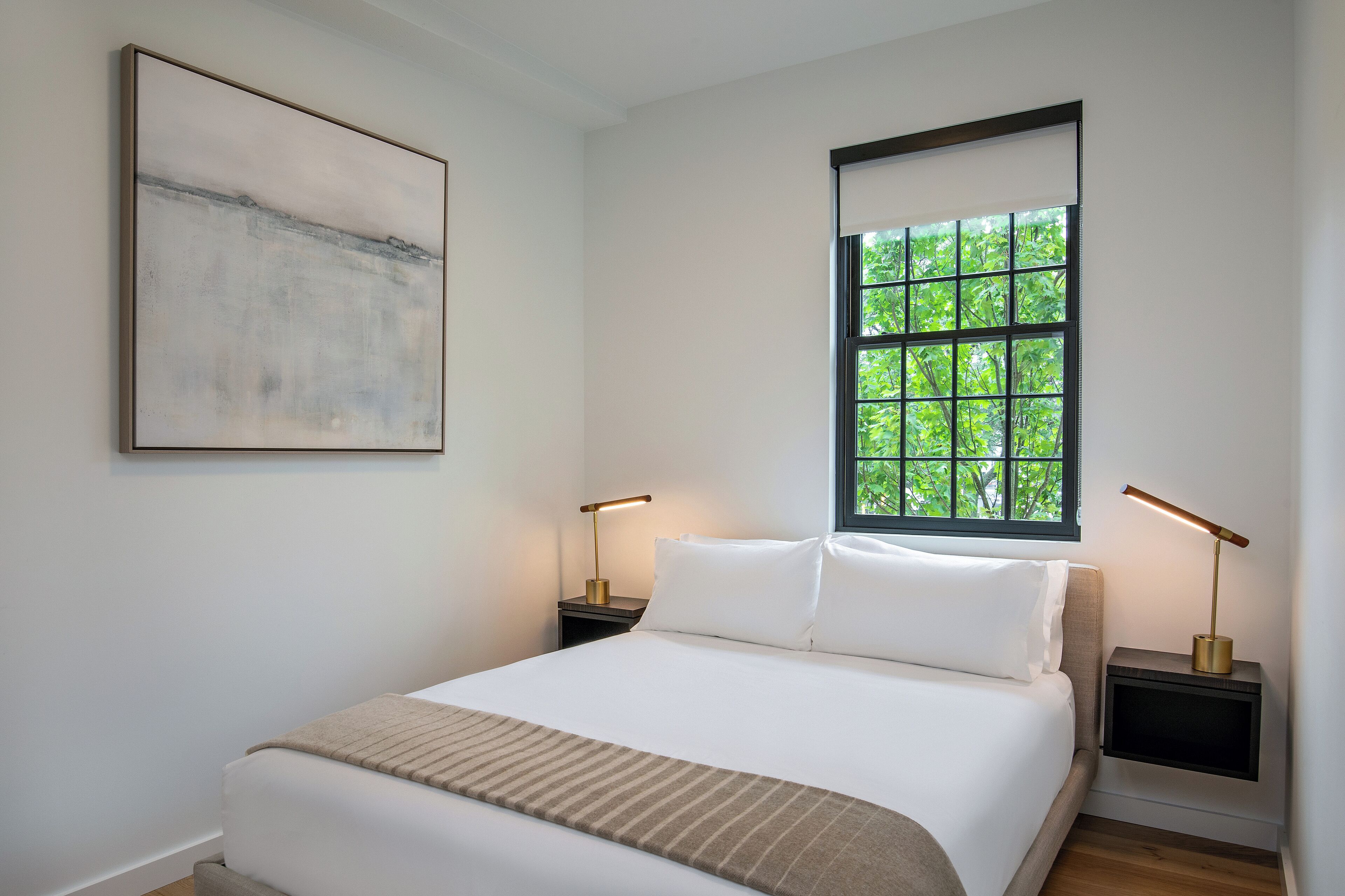 Een netjes opgemaakt bed met witte lakens, twee nachtkastjes met lampen, een groot abstract schilderij aan de muur, en een raam met uitzicht op groen.