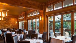 Een restaurant met houten wanden en grote ramen, tafels gedekt met witte tafellakens en stoelen.