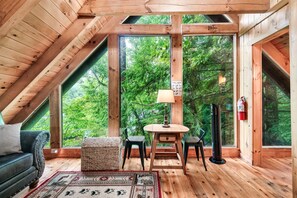 A cozy attic room with wooden beams, a large window overlooking a forest, a sofa, a table, and a chair. - Free Dollywood Tix/Pool/Arcade/HotTub/A+Location (Gatlinburg)