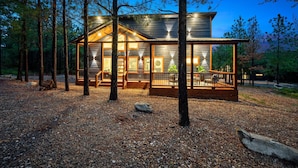 A wooden cabin with large windows and a deck, surrounded by trees. - Sauna, Bar, Big Patio, Hot Tub, Fire Pit & Pets (Broken Bow)