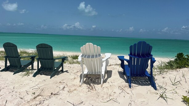 Tres sillas Adirondack mirando a una playa con agua turquesa cristalina y cielo despejado.
