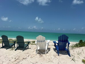 Trois chaises Adirondack sont disposées face à une plage, avec une eau turquoise cristalline et un ciel dégagé.