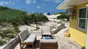 Un patio con dos sillones de madera y una mesa pequeña, con vista a una playa arenosa con vegetación y un edificio distante.