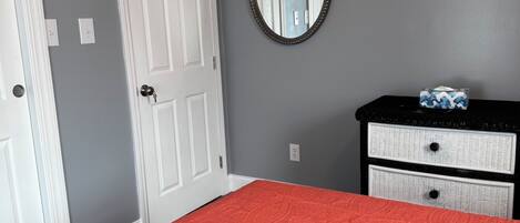 A bedroom with a red bedspread, a white door, a dresser with two drawers, and a round mirror on the wall.