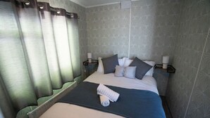 A neatly made bed with a blue blanket, pillows, and a rolled towel. There are two bedside tables with lamps. The room has patterned wallpaper and a curtain. - No.16 Nursery park caravan site (Brechin)