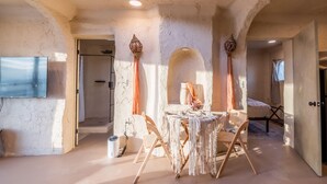 Een kamer met een rustieke stenen muur, een houten tafel met macramé tafelkleed, en een bed met een wit laken.