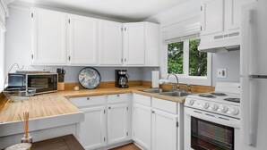 A modern kitchen with white cabinetry, a wooden countertop, a built-in stove, and a window with a view of greenery. - Relax at Hilltop Hideaway, Near the Shore (Dennis Port)