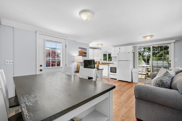 Una cucina moderna con mobili bianchi, un'area pranzo con piano di lavoro scuro e un soggiorno con divano e cuscini decorati.
