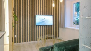 A modern room with a striped wall, a flat-screen TV, two glass tables, a green sofa, and a window with a view of greenery.