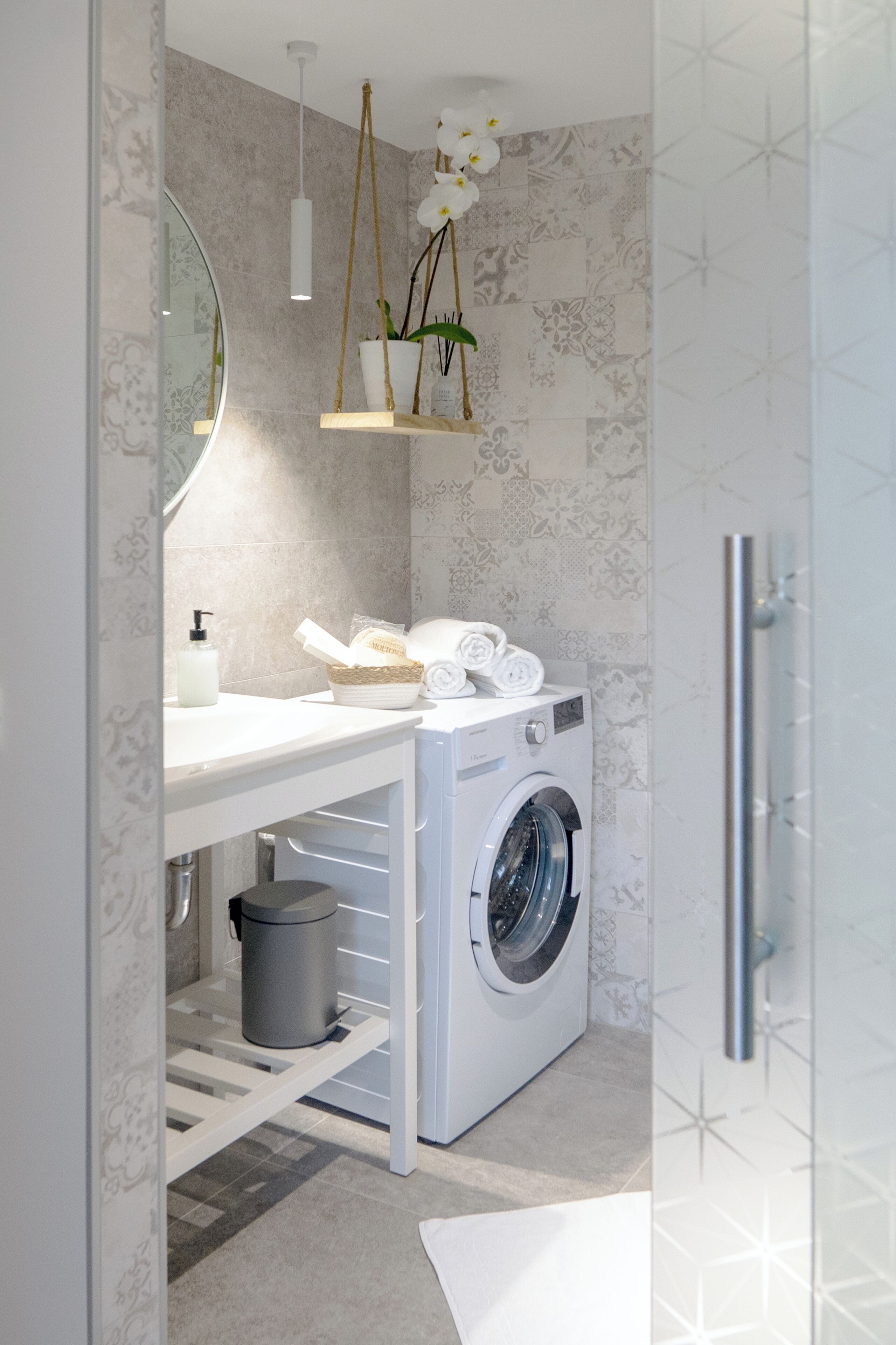 Ein kleines, modernes Badezimmer mit Waschmaschine, Waschbecken mit Spiegel und einer hängenden Orchideenpflanze.