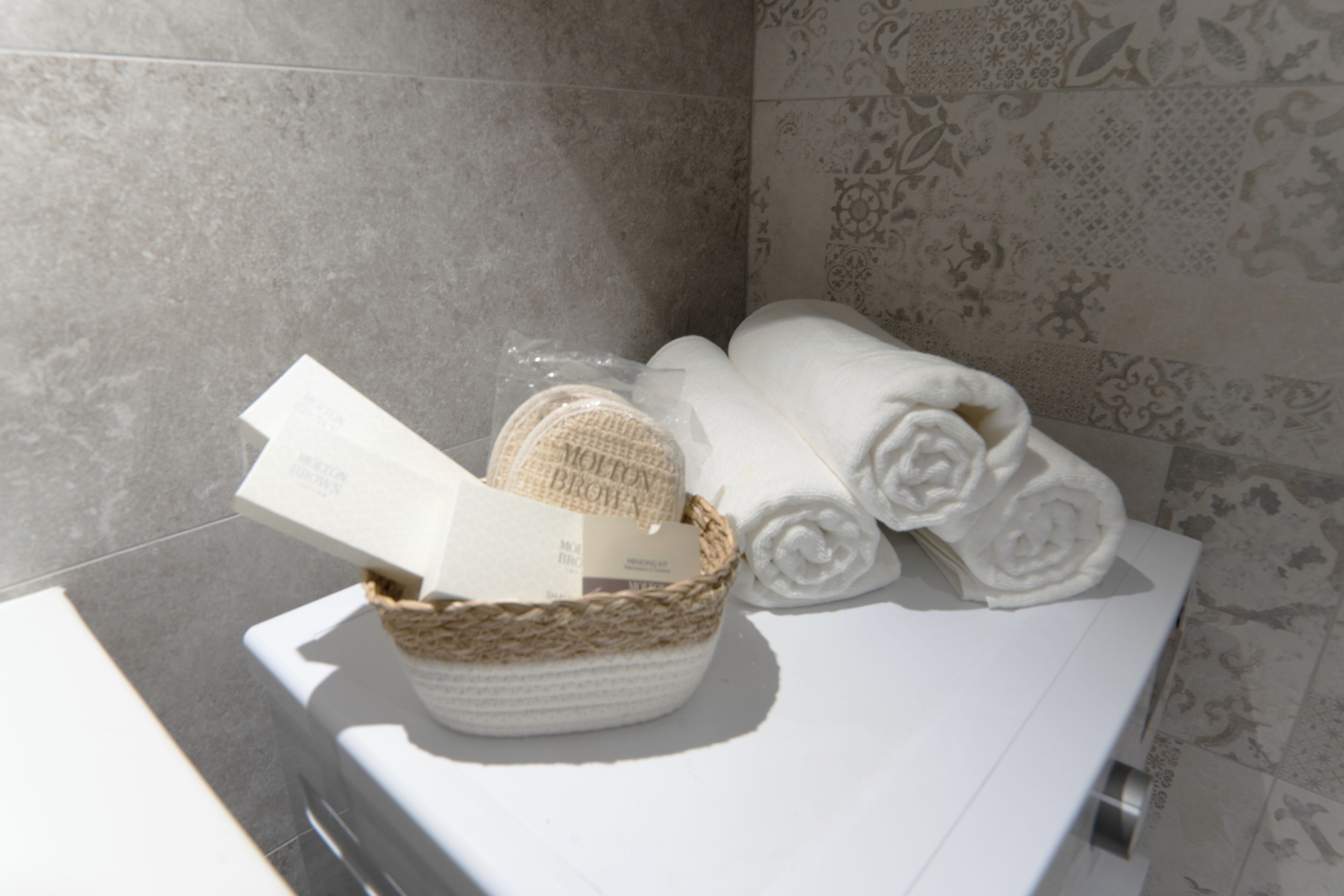 Een badkamer met een witte wastafel, een gevlochten mand met toiletartikelen, en netjes opgerolde handdoeken.