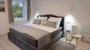 A modern bedroom with a large bed, a small desk, and a view of greenery outside.